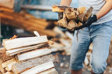 Tuinposter Een stapel gestapeld brandhout, voorbereid voor het verwarmen van het huis. Brandhout verzamelen voor de winter of een kampvuur. Man houdt brandhout in handen © Anastassiya 