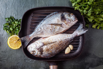 Two raw dorado fish with spices, lemon and parsley in black grill pan on black concrete background....