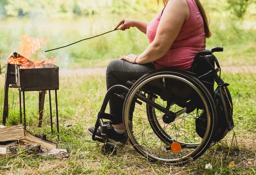 Disabled Woman Fries Meat At The Stake In A Campsite With Friends. Wheelchair In The Forest On The Background Of Bonfire. Barbeque. Camping.