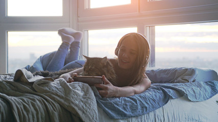 Young woman in headphones relaxinglisten to music laing down in bed by the window takes selfie with her lovely big Maine Coon cat during sunset with lens flare effects