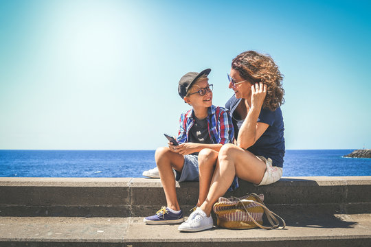Smiling mother and son watching smartphone together sitting near the sea. Woman and trendy young boy playing with cell phone. Happy family enjoying summer holiday. Happiness, togetherness concept.