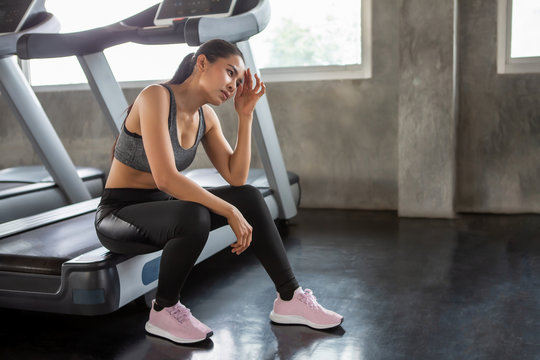 Beautiful Asian Young Woman Tired Taking A Break From Running Or Exercise Sitting On Treadmill Machine  In Fitness Gym Healthy .girl In Sportswear Workout Rest In Morning