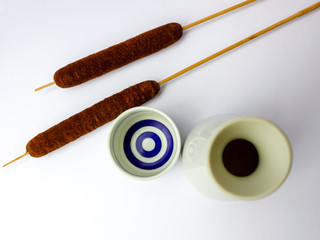 white sake set from a bottle and a cup with blue stripes and cattail on white backdrop view from up