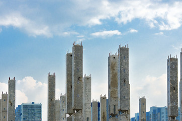 Fototapeta na wymiar Vertical Reinforced Concrete Columns with Rebars at Construction Site, Finished Apartment Buildings, Sky and Clouds in Background. Real Estate, Residential Buildings Urban Mixed-Use Development.