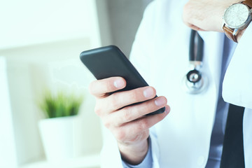 Male doctor in white coat is using a modern smartphone device with touch screen. Doctor hands with mobile phone.