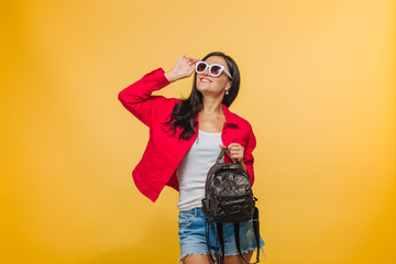 Girl in sunglasses on yellow background in red jacket and shorts with black backpack