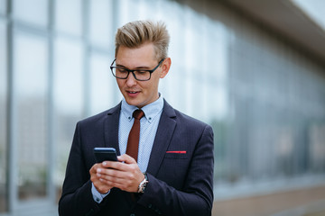 Happy businessman smiling as he reads a text message standing outside the office. Male executive manager in trendy clothes using smartphone outdoors. Modern technology concept