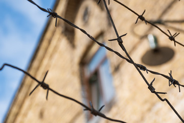 Close-up of a rusty barbed wire fence surrounding a concentration and extermination camp, old barrack with window and lamp in soft focus in the background