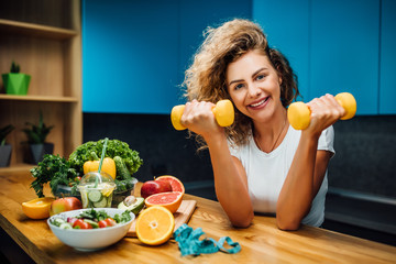 Portrait of fit and strong girl, model and fitness trainer holding dumbbells, with fresh salade on table, healthy eat time.