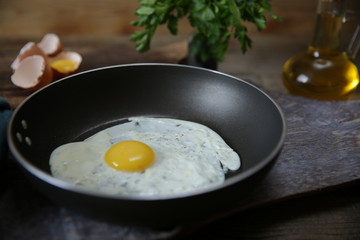 Close-up. Frying pan with frying egg with yolk on a wooden cutting board with ingredients for the preparation of fried eggs: raw eggs, olive oil, salt, parsley. 