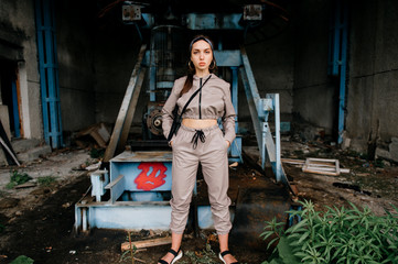 Beautiful young stylish girl posing at abandoned factory outdoor with an old broken engine.