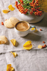 Obraz na płótnie Canvas Cup of espresso coffee standing on linen table cloth with yellow autumn leaves, berries in ceramic vase and shortbread cookies with dark wall at background. Fall concept. Copy space