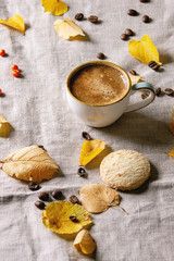 Cup of espresso coffee standing on linen table cloth with yellow autumn leaves, berries, coffee beans and shortbread cookies with dark wall at background. Fall concept. Copy space