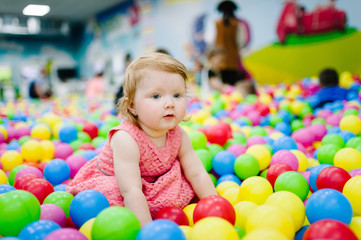 Fototapeta na wymiar Happy laughing girl playing with toys, colorful balls in playground, ball pit, dry pool. Little cute child having fun in ball pit on birthday party in kids amusement park and play center indoor.