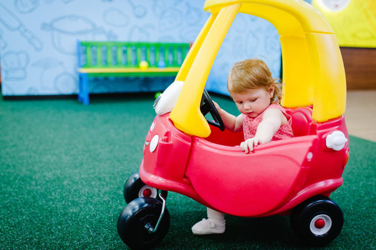 Children, little girl child rides on a red big car on the road. Close up. The baby is driving the car and played in a children's playroom on a birthday party. Time together at entertainment centre.