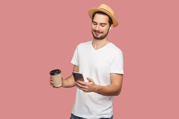 Portrait of satisfied handsome bearded young hipster blogger man in white shirt and casual hat standing with coffee and chatting on the phone. Indoor, isolated, studio shot, pink background