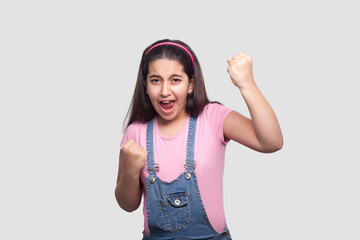 Obraz na płótnie Canvas Portrait of rejoicing winner brunette young girl in casual pink t-shirt and blue denim overalls standing, screaming and celebrating her victory. indoor studio shot, isolated on light gray background.