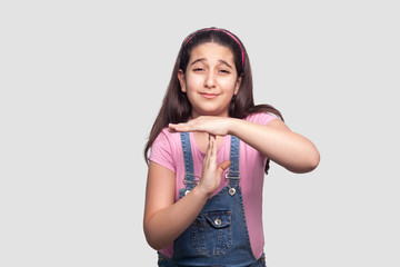 I need more time. Portrait of hopeful sad brunette young girl in pink t-shirt and blue overalls standing with timeout sign and asking for more. indoor studio shot, isolated on light gray background.