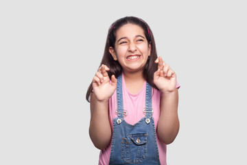 Portrait of hopeful beautiful brunette young girl in casual style, pink t-shirt and blue denim overalls standing with crossed fingers and clenching teeth. studio shot isolated on light gray background