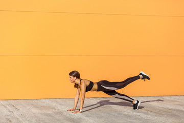 Young satisfied beautiful woman wearing black sporwear practicing sport exercises in morning on street, standing on plank position with raised leg, holding balance, orange wall background, outdoor