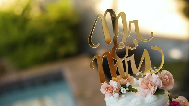 Shot of a beautifully decorated floral wedding cake sitting on a pedestal right next to a pool during a summer wedding reception.