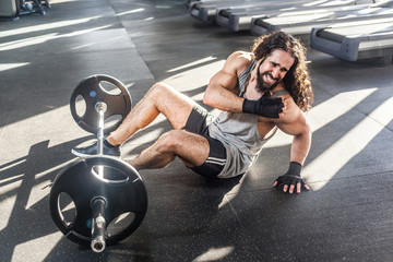 Portrait of grimace young adult man athlete with long curly hair working out in gym, sitting on...