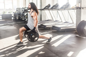 Side view of concentration young adult sportman athlete with long curly hair working out in gym, squating on one knee and holding two disks, doing exercises for legs, squatting. indoor, looking away