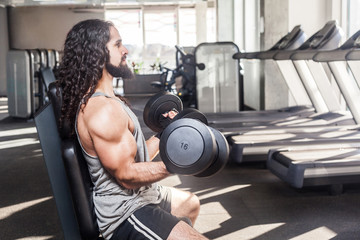 Side view portrait of concentration young adult man with long curly hair working out in gym, sitting on bench and holding two dumbbell with raised arms, doing exercises for biceps. indoor,looking away