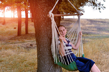 The girl in the park is resting and sitting on a hammock, against the background of the evening sun and looking into the distance.