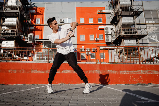 Young guy dressed in jeans and t-shirt is dancing modern dance in the street on the background of urban buildings in the warm day