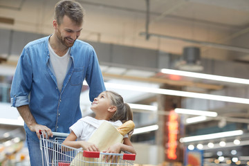 Portrait of happy young father grocery shopping in supermarket and smiling at little girl sitting...