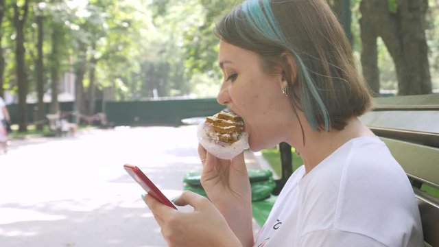 Charming woman in white T-shirt reads message on red smartphone and eating piece of cake with summer park background. Break time concept