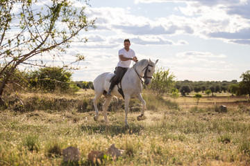 Young guy in casual outfit riding white horse on meadow a sunny day 
