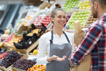 Waist up portrait of pretty young saleswoman talking to customer while standing by fresh fruits and...