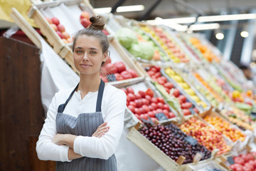 Waist up portrait of pretty young saleswoman looking at camera while posing by fresh fruits and...