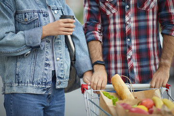 Mid-section portrait of contemporary young couple pushing shopping cart with groceries in parking...