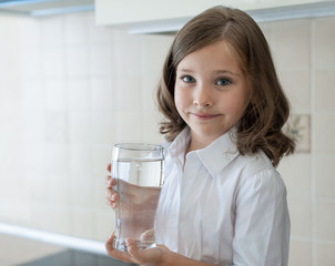 Little child is drinking clean water at home, close up. Caucasian cute girl with long hair is holding a water glass in her hands. Taking care of own health. Concept of healthy lifestyle, good habit