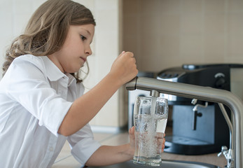 Portrait of a little caucasian girl gaining a glass of tap clean water. Kitchen faucet. Cute curly kid pouring fresh water from filter tap. Indoors. Healthy life concept