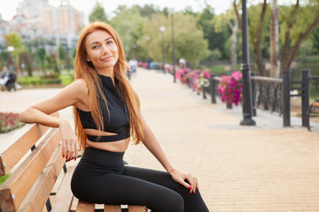 Happy beautiful fitness woman smiling to the camera, sitting on the bench outdoors after morning workout, copy space. Lovely sportswoman resting outdoors after working out