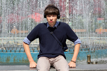 A young man, a teenager, sits at a fountain in a city park and listens to music in headphones.