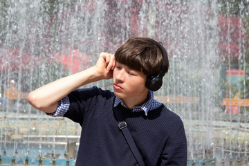 A young man, a teenager, sits at a fountain in a city park and listens to music in headphones.