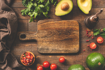 Fresh vegetables and cutting board for cooking, food frame background. Avocado, tomato, salsa, parsley and pepper spices on wooden table