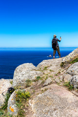 Obraz premium Male pilgrim on the rocks at Cape Finisterre, Cabo Fisterra - the end of the Way of St. James takes pictures with digital compact camera