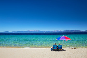 Couple sitting under umbrella enjoying the turquoise waters and expansive view of Lake Tahoe-...