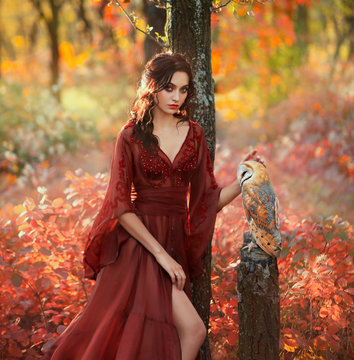 Fiery Bloody Lady In A Long Light Summer Red Burgundy Dress With An Open Leg, Girl With Black Hair In A Gothic Style Stands In Forest And Strokes A Cute White Barn Owl, Mythical Forest Creatures