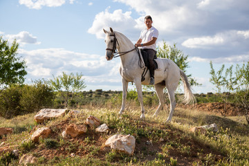 Young male in casual outfit riding white horse on grassy meadow a sunny day