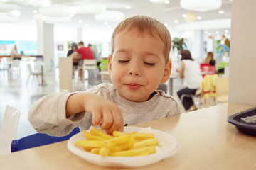 little child eating french fries sitting at a table on the food court of the mall. Unhealthy food.