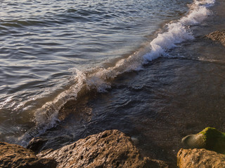 Sea waves beat against coastal sand and stones at sunset on a summer sunny evening.