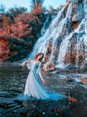 lovely girl with fair tender skin stands in aqua in long light blue dress, mystical mermaid near high waterfall with powerful streams of cold water, Lost doll with Korean face and waterproof makeup