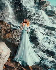 sweet lady in light long blue flying dress, water nymph stands near large waterfall in spray of...